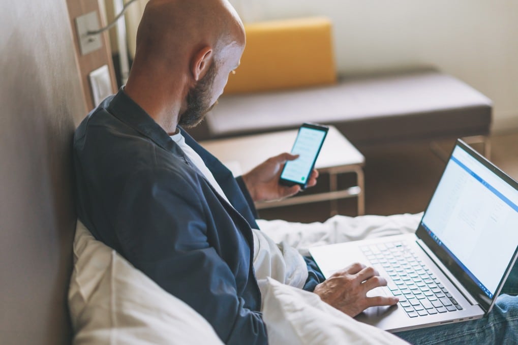 adult-bald-bearded-man-in-blue-jacket-with-mobile-phone-working-on-laptop-in-hotel-room - kleesto tour operator software analytics